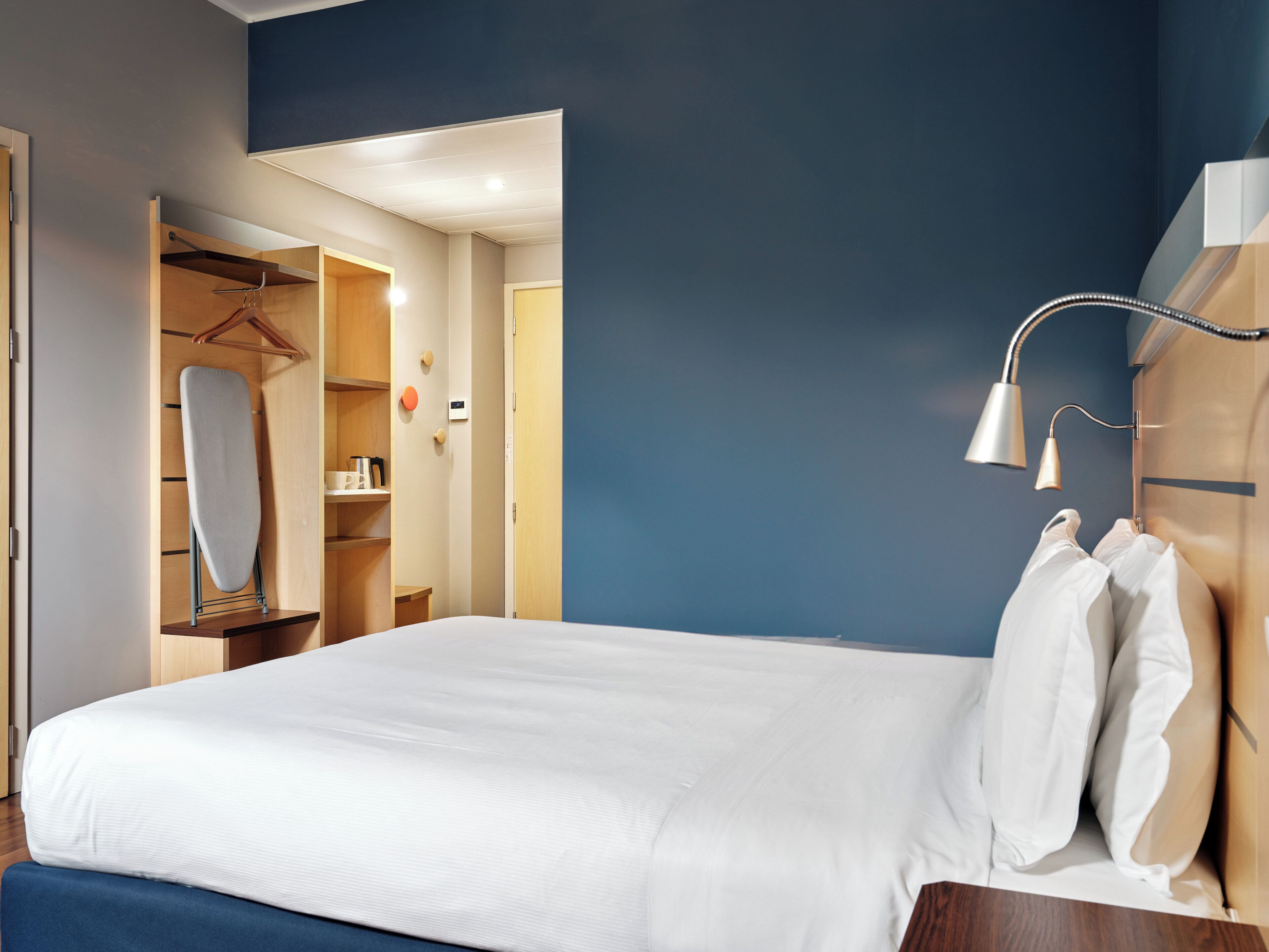 Une chambre d’hôtel avec un lit bien fait, une armoire, un vêtement suspendu et une lampe de chevet.