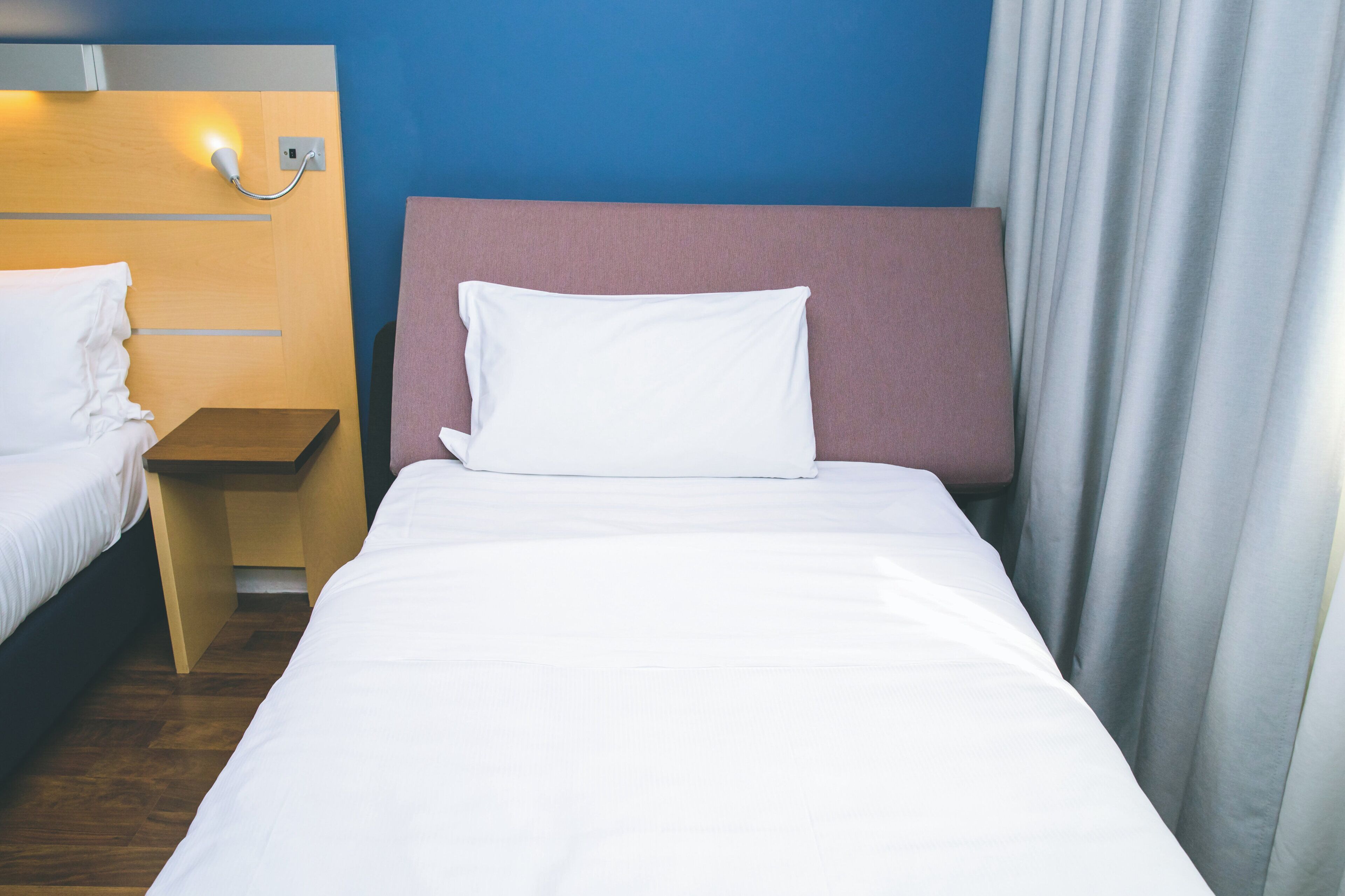 Habitación de hotel con una cama individual, mesita de noche, lámpara de pared y ventana con cortinas.