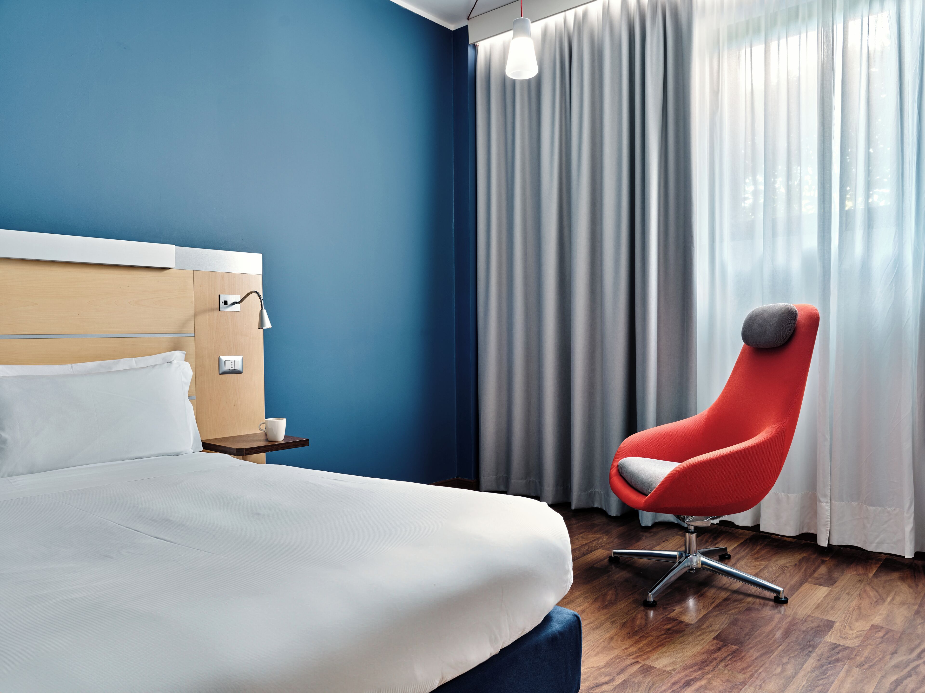 A hotel room with a bed, a red chair, a nightstand, and a window with curtains.