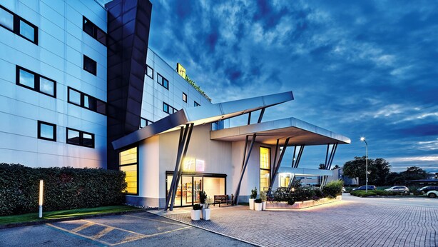 A modern hotel with a glass facade, large windows, and a prominent entrance with a covered area. - Holiday Inn Express Milan - Malpensa Airport by IHG (Somma Lombardo)
