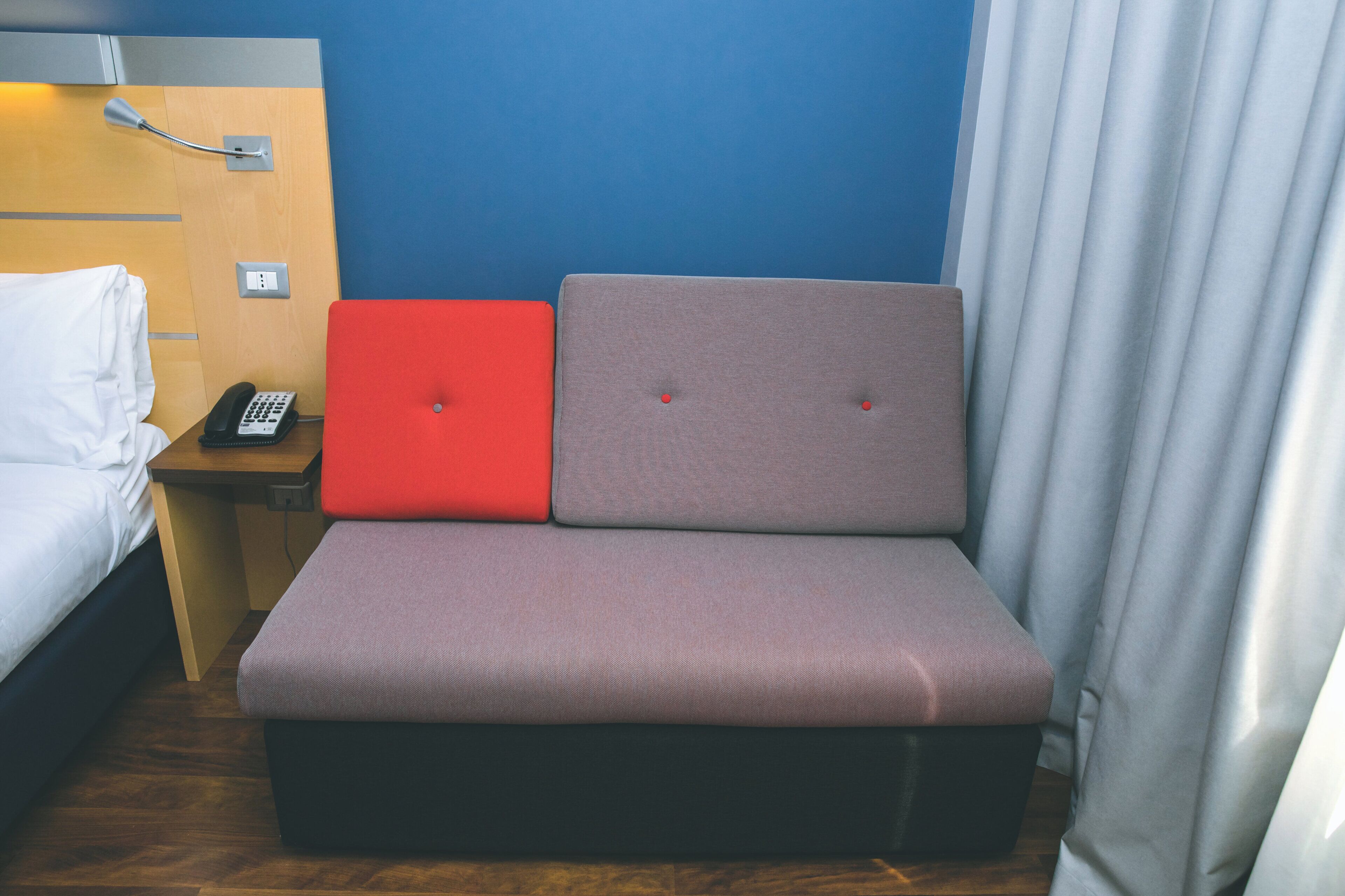 Quarto de hotel compacto com cama, mesa de cabeceira pequena com telefone e área de estar com um banco estofado em vermelho e cinza.