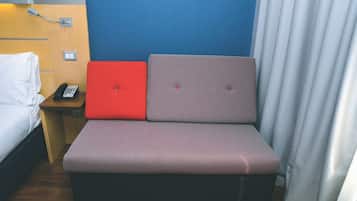 Quarto de hotel compacto com cama, mesa de cabeceira pequena com telefone e área de estar com um banco estofado em vermelho e cinza.