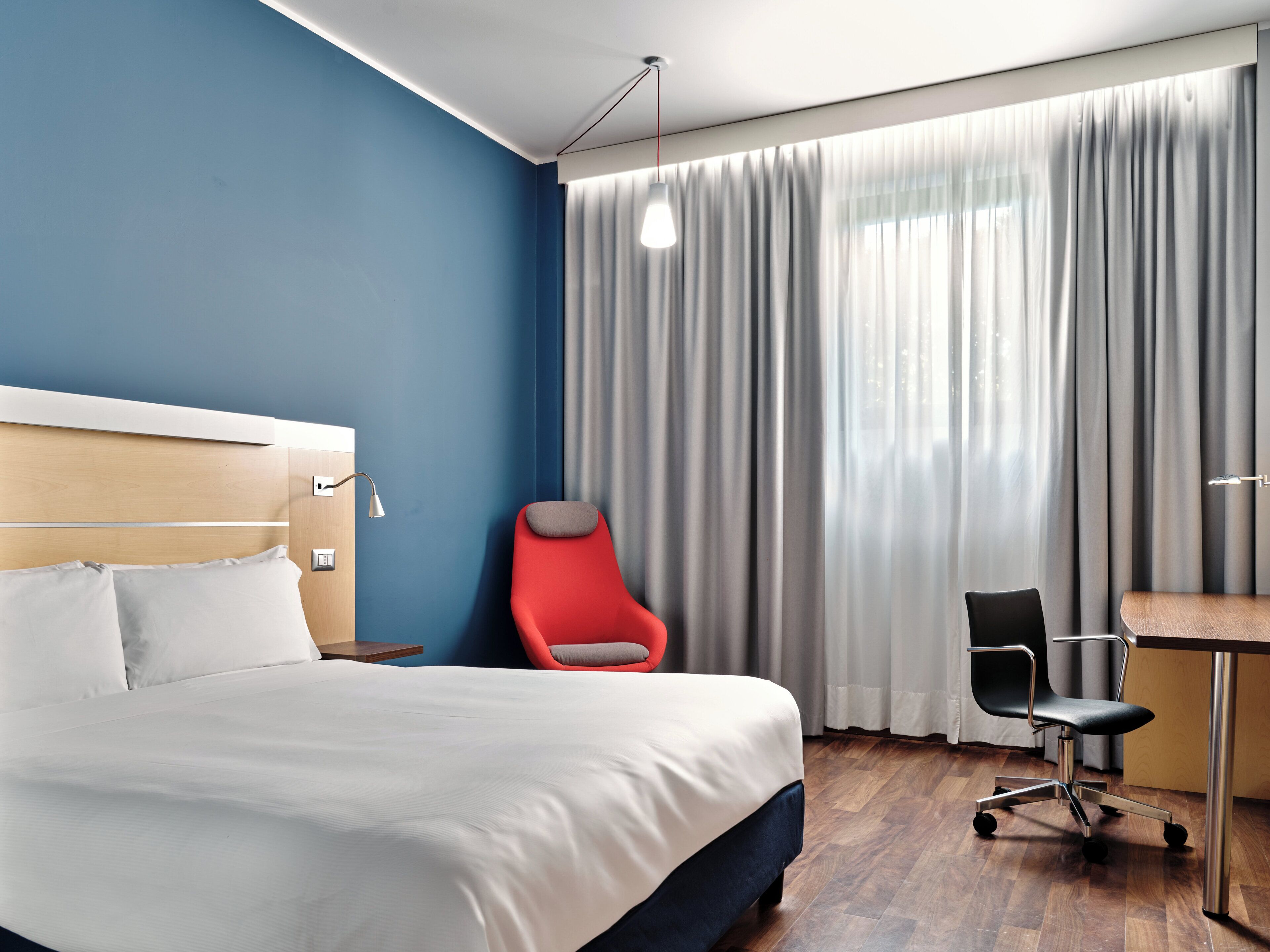 A hotel room with a bed, a desk, a chair, and a large window with curtains.