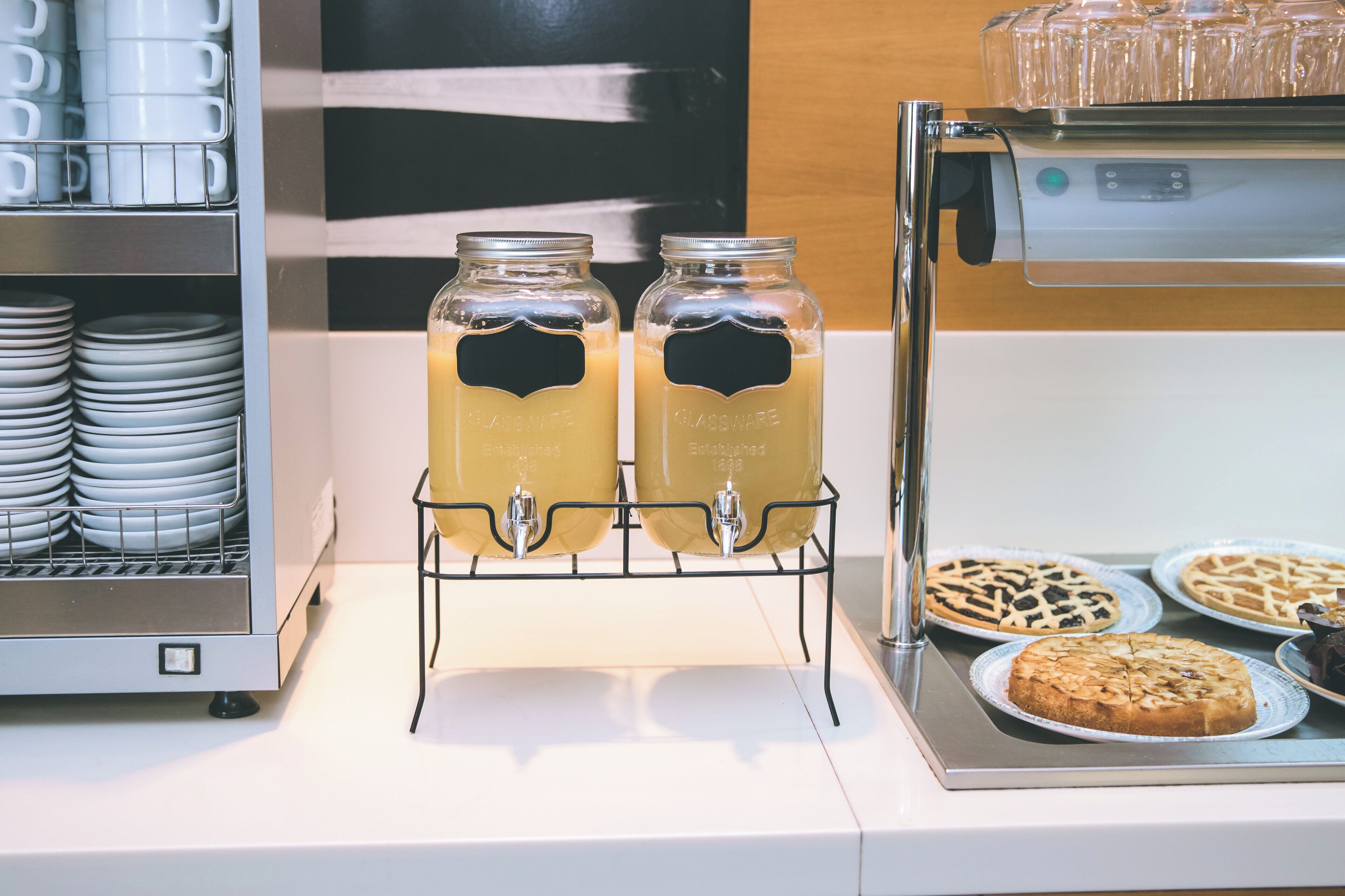 Un área de bebidas con dos frascos de vidrio sobre un soporte metálico, una cafetera y una selección de platos y pasteles.