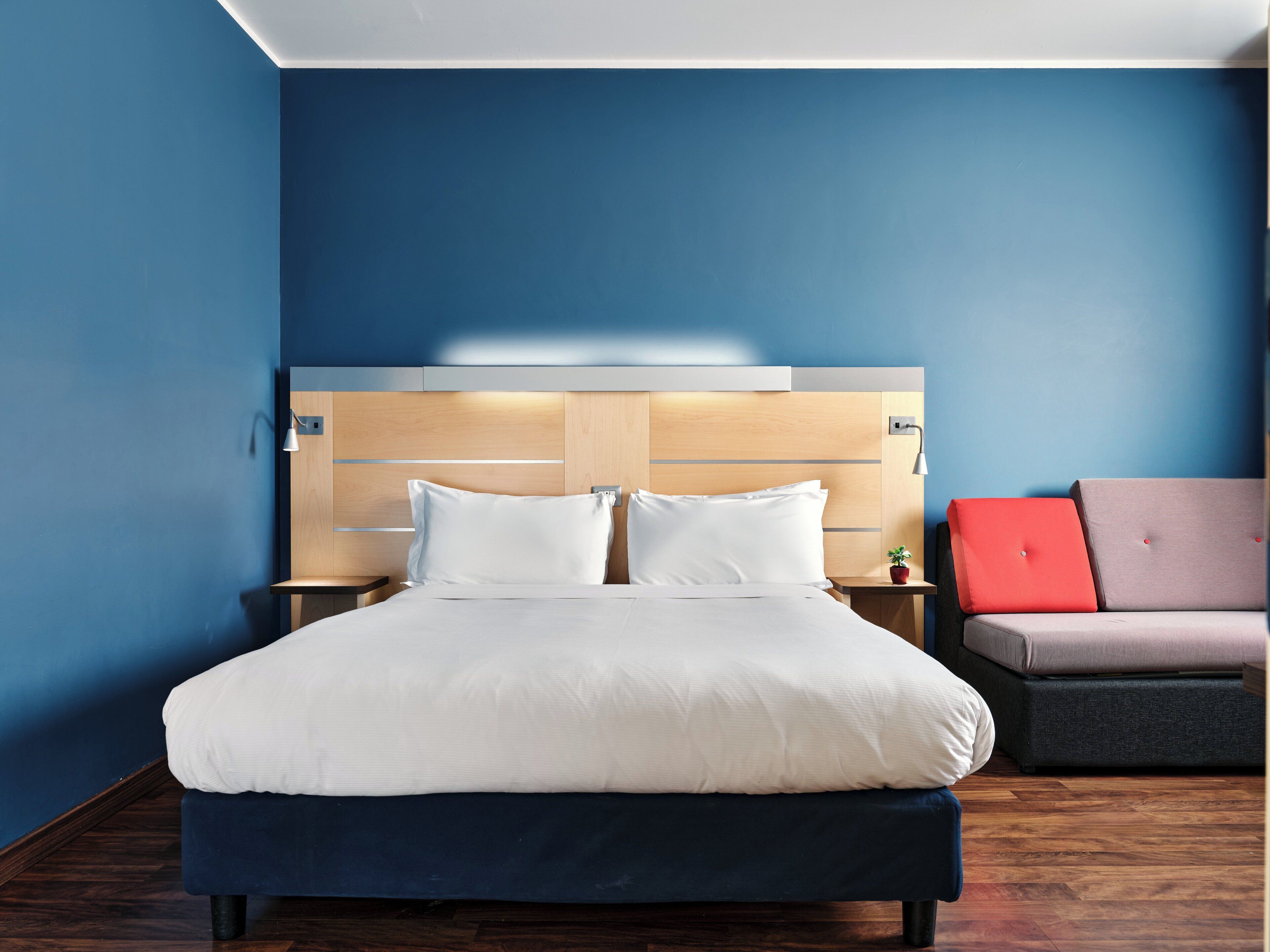 A hotel room with a bed, nightstands, a red and grey sofa, and a blue wall.