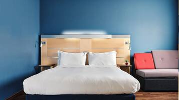 A hotel room with a bed, nightstands, a red and grey sofa, and a blue wall.