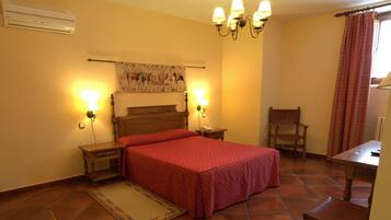Un dormitorio con una cama de madera, dos mesitas de noche, un aparador, una silla, una mesa, un aire acondicionado instalado en la pared, una araña y un tapiz.