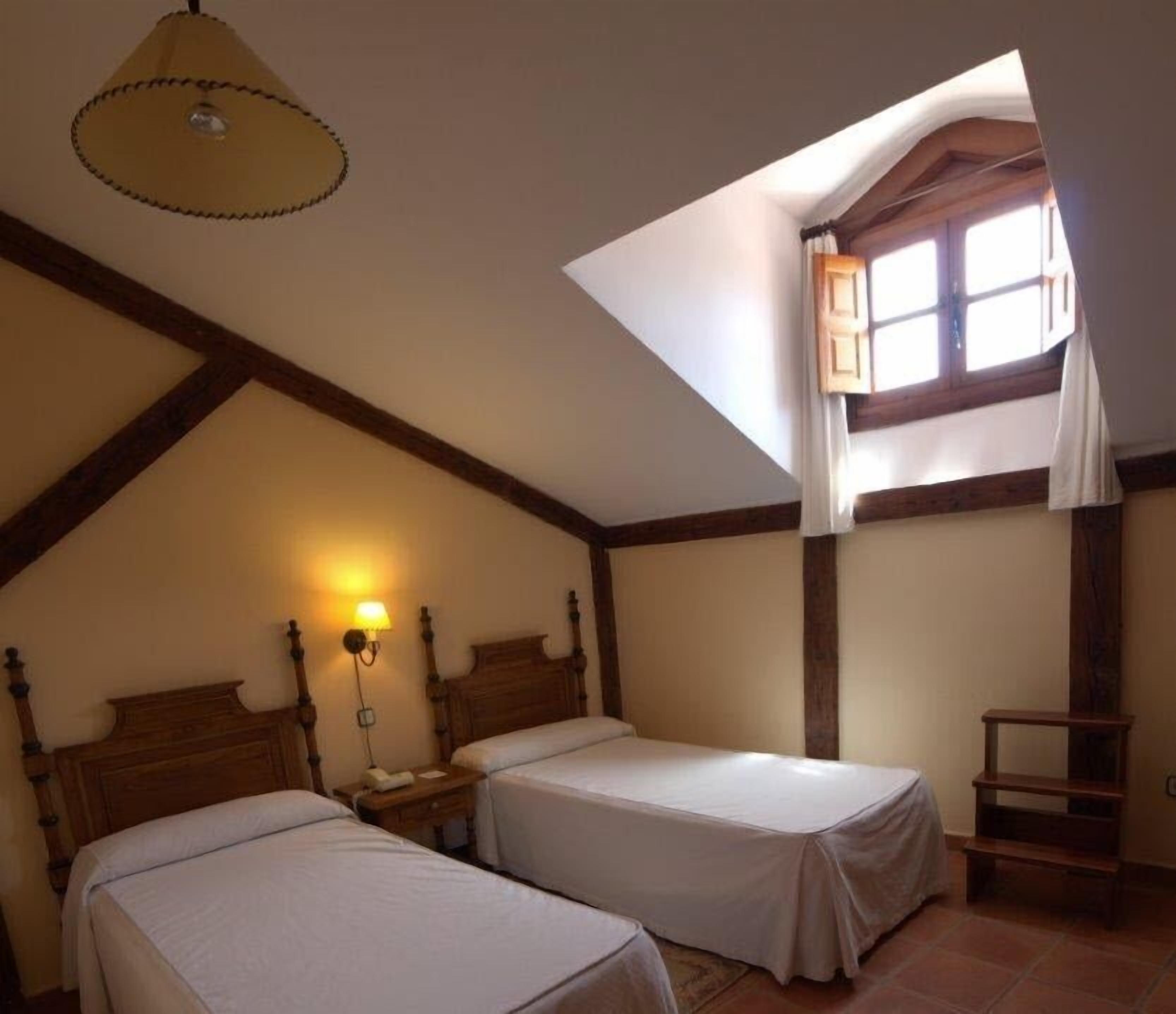 Una habitación con dos camas individuales, techo inclinado, tragaluz y un pequeño estante de madera.