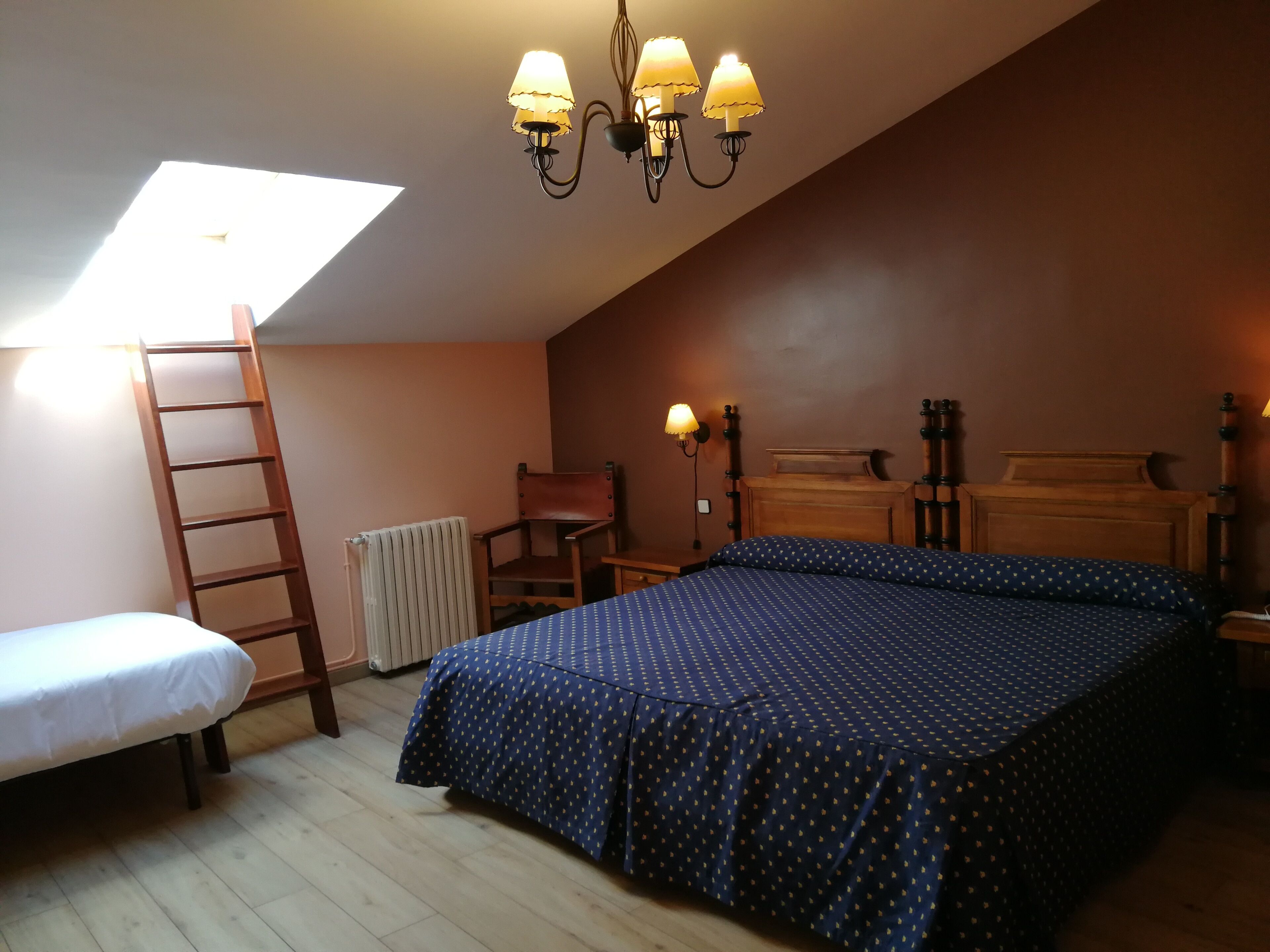 Un dormitorio con techo inclinado, una cama con colcha estampada, una silla de madera, una mesita de noche de madera con lámpara y una escalera apoyada en la pared.