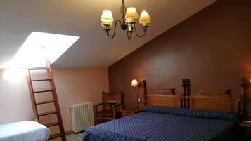 Un dormitorio con techo inclinado, una cama con colcha estampada, una silla de madera, una mesita de noche de madera con lámpara y una escalera apoyada en la pared.