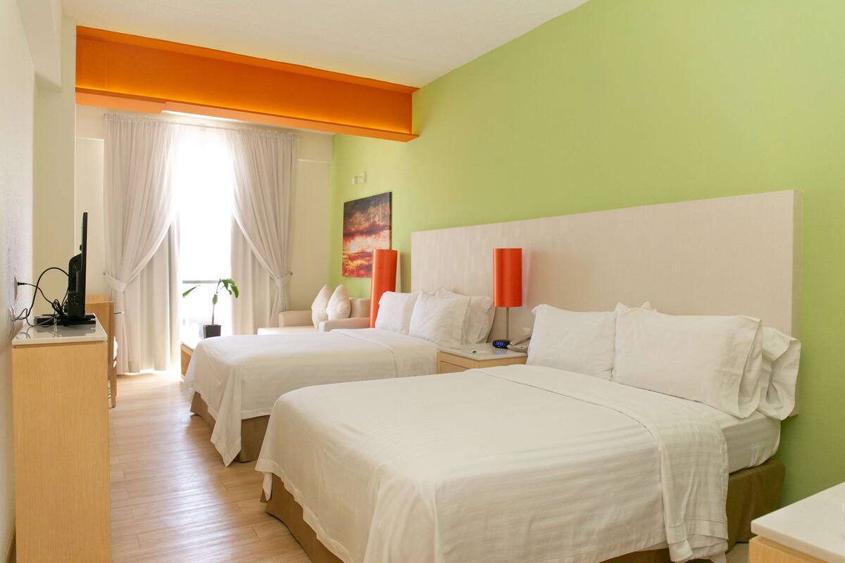A hotel room with two beds, a desk, a TV, and a window with curtains.
