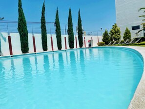 Una piscina con superficie de agua azul cristalina, rodeada de árboles altos y delgados y un edificio de fachada blanca con detalles rojos.