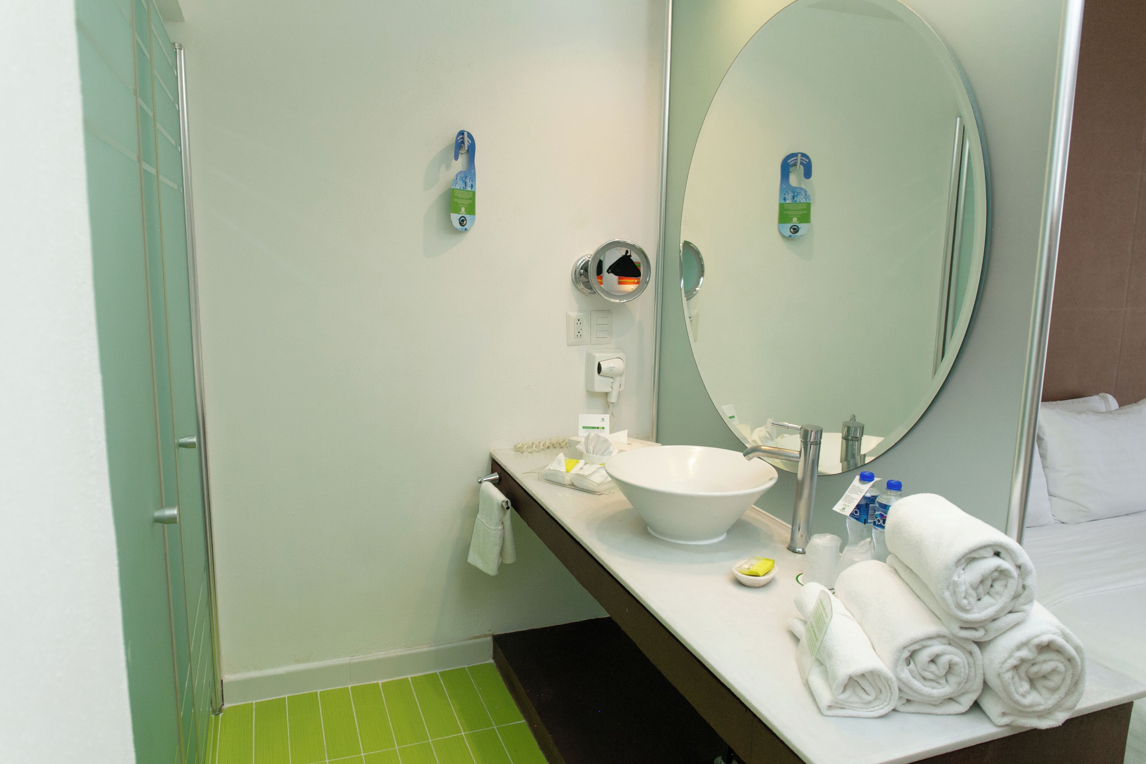 A hotel bathroom with a white sink, a large mirror, a towel rack, and a green-tiled floor.