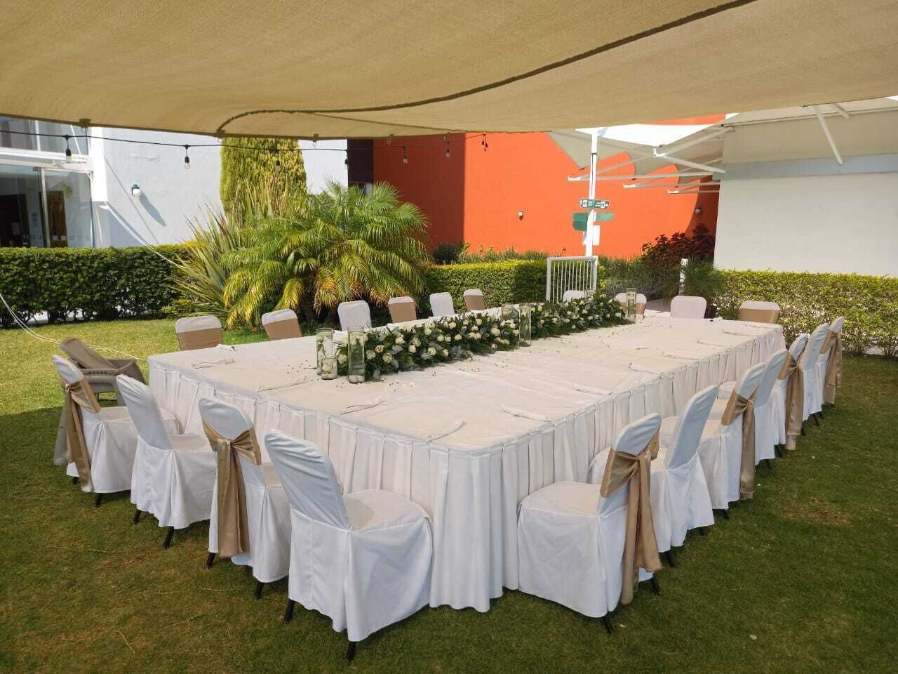 Une table rectangulaire installée à l’extérieur, sous une tonnelle, entourée de chaises recouvertes d’housse blanche et beige, et agrémentée d’un arrangement floral au centre.