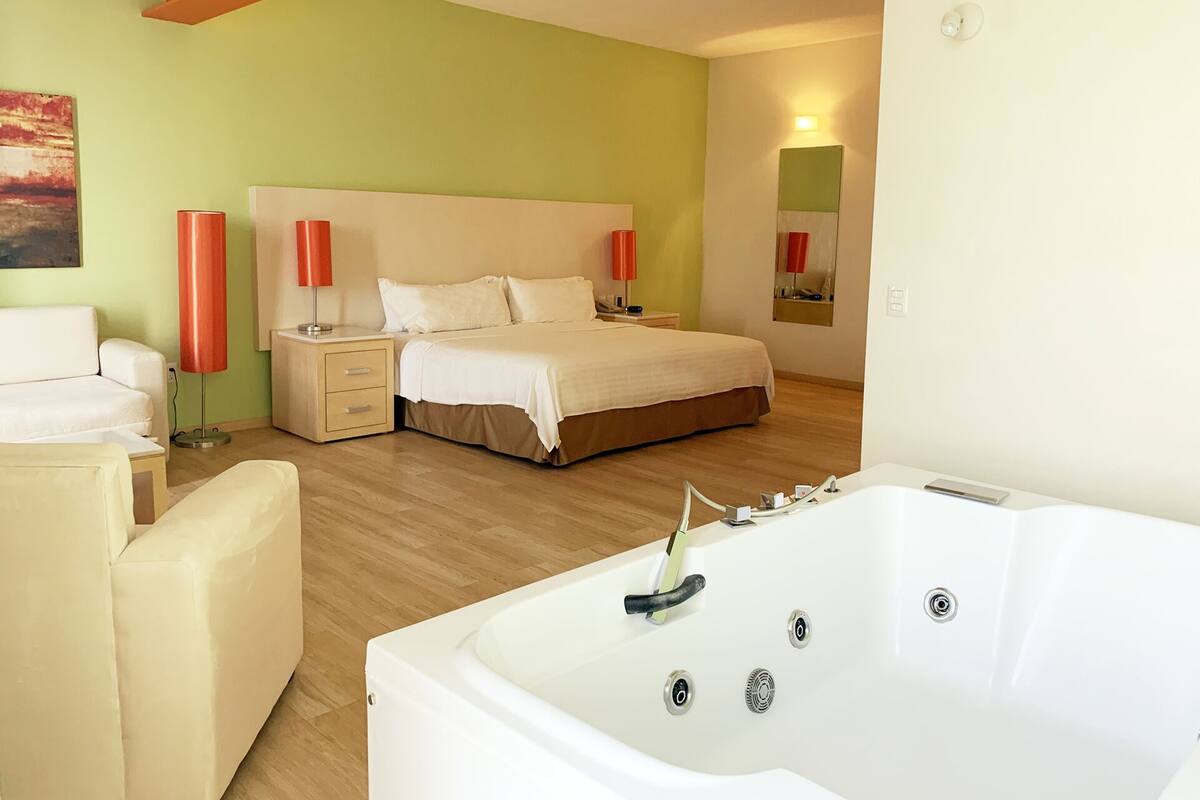 A modern hotel room with a large bed, a sitting area with a chair, a large bathtub, and a nightstand with a lamp.