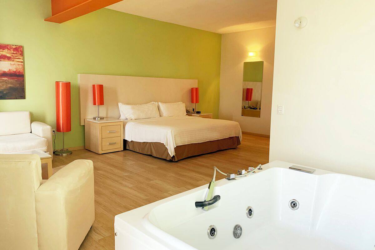 A hotel room with a bed, bedside tables, a chair, a large bathtub, and a mirror on the wall.