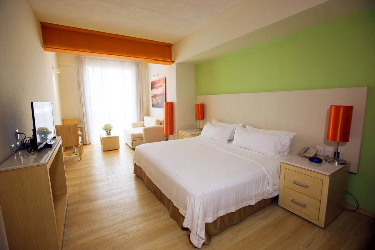 A hotel room with a large bed, bedside tables, a desk with a computer, a chair, and a window with curtains.