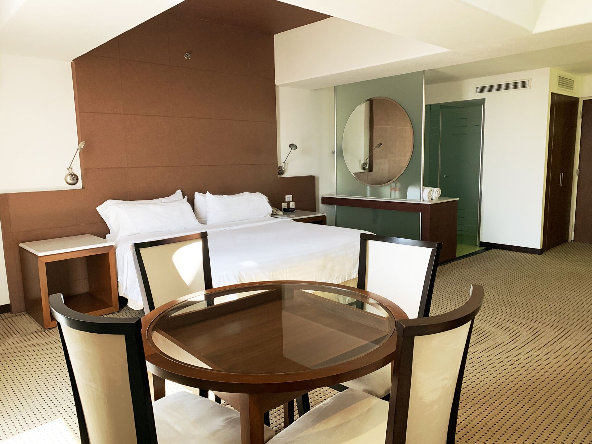 Habitación de hotel con cama, mesitas de noche, una mesa redonda de vidrio con sillas, un espejo grande y un armario.