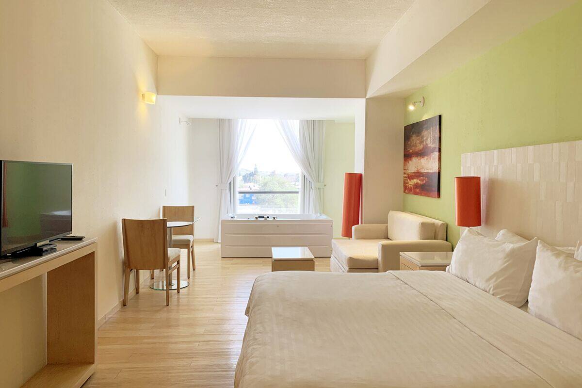 A hotel room with a bed, a desk with a TV, a sofa, a chair, and a window with curtains.