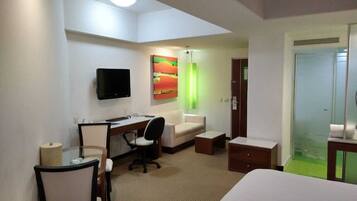 Habitación de hotel con cama, escritorio, silla, televisor y ducha de vidrio.