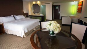 Habitación de hotel con una cama grande, una mesa redonda con un florero, un espejo y una zona de estar con sillas.
