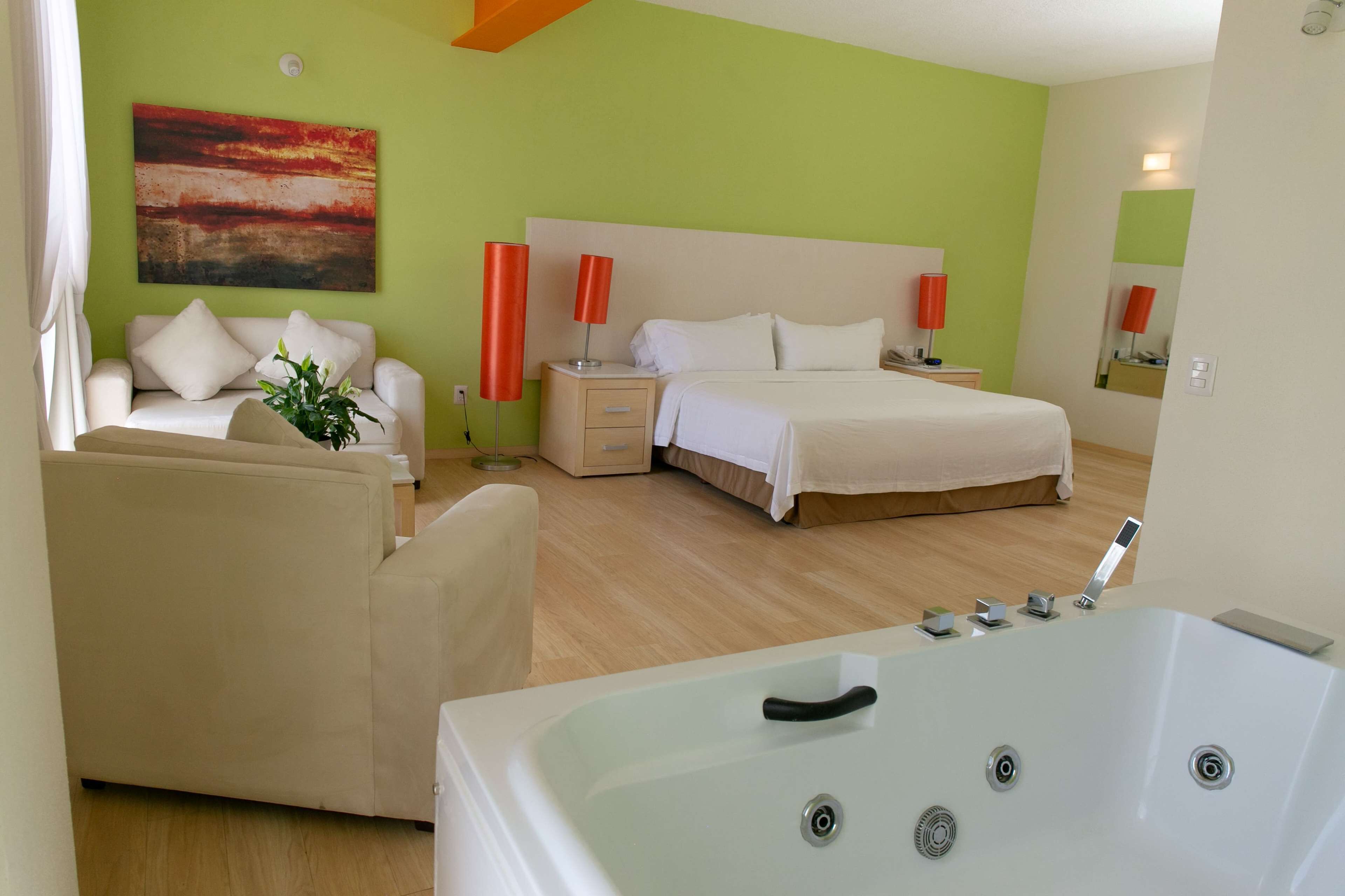 A hotel room with a bed, a sofa, a nightstand, a red lamp, a large window, and a bathroom with a bathtub.