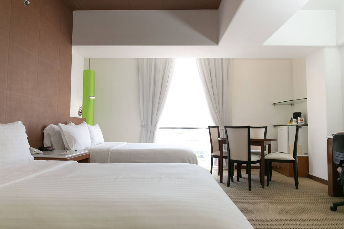 A hotel room with two beds, a desk, chairs, and a bright window.
