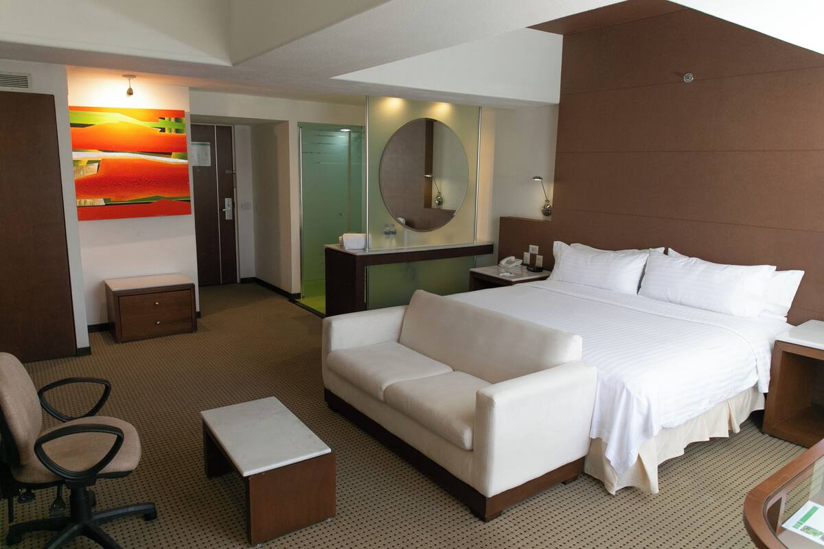 A hotel room with a bed, a sofa, a desk, a chair, a mirror, and a painting on the wall.
