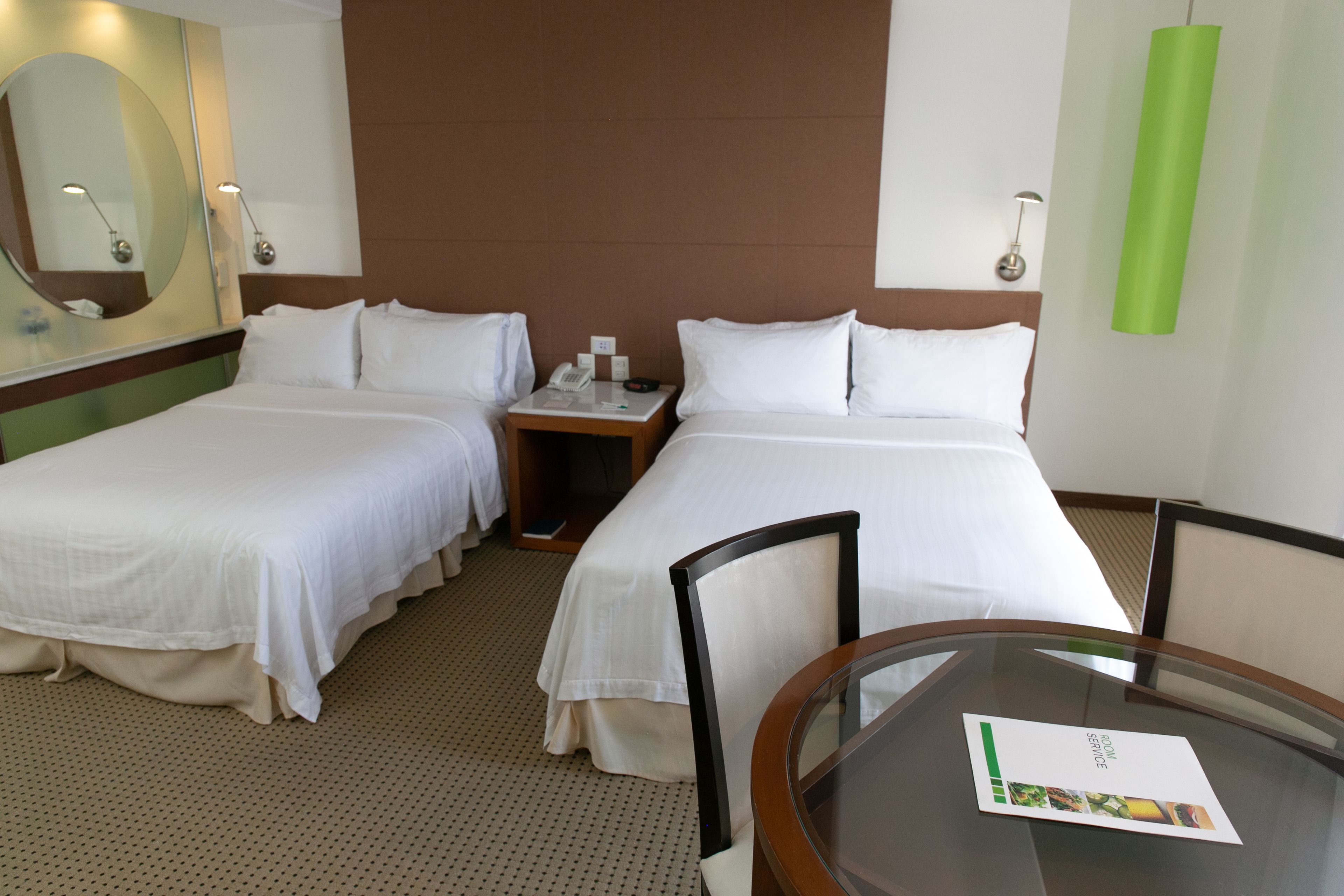 A hotel room with two single beds, a small table with a menu, a mirror, and a green pendant light.