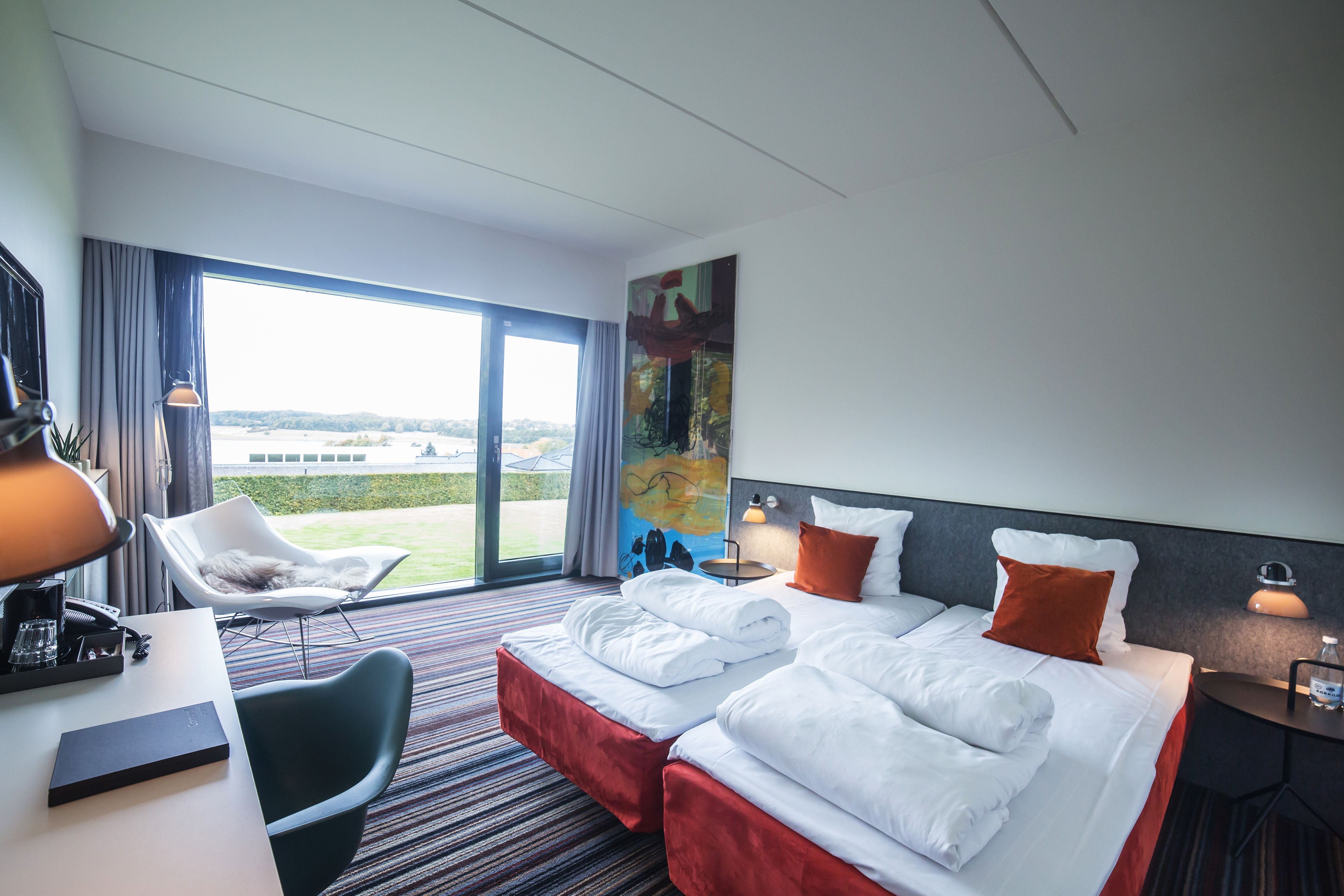 Une chambre d’hôtel avec un grand lit, deux tables de chevet, un bureau avec une lampe, une chaise et une vue sur l’extérieur.