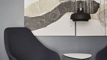 A modern room with a grey wall, a large abstract painting, a black pendant light, and two dark blue chairs with a small white table.