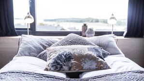 A bed with a patterned blanket and pillows, a view of a body of water through a window, and two bedside lamps. - Comwell Roskilde (Roskilde)