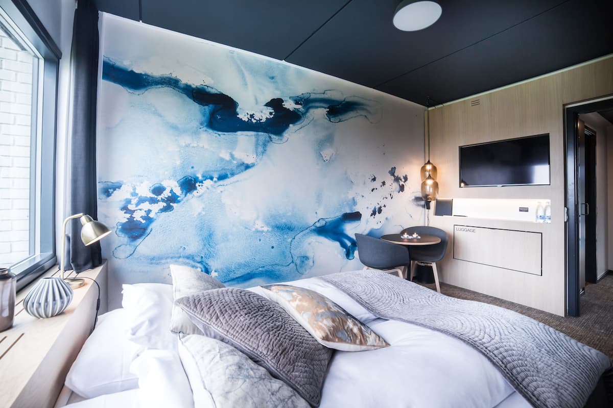 Moderni hotellihuone, jossa on suuri, abstrakti taulu seinällä, useilla tyynyillä varustettu sänky, työpöytä tuolilla, taulutelevisio ja matkatavaroiden säilytystila.
