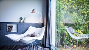 A modern room with a white chair, a floor lamp, and a view of trees through a large window. - Comwell Roskilde (Roskilde)