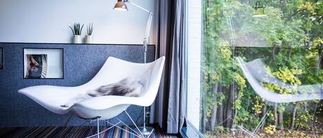 Una habitación moderna con una silla blanca, una lámpara de pie y vistas a árboles a través de un ventanal amplio.