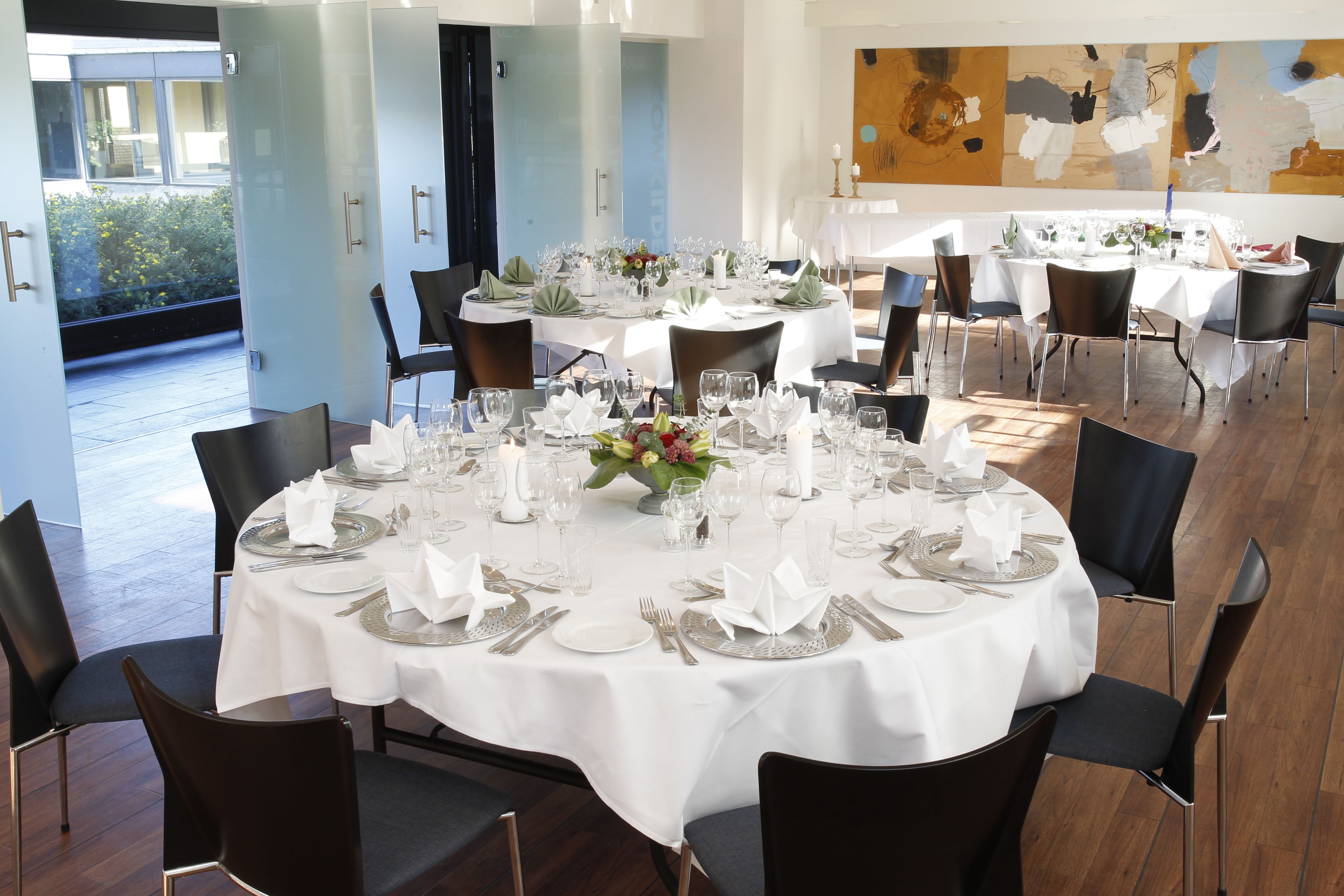 A dining area with round tables set for a meal, featuring white tablecloths, silverware, and glassware.