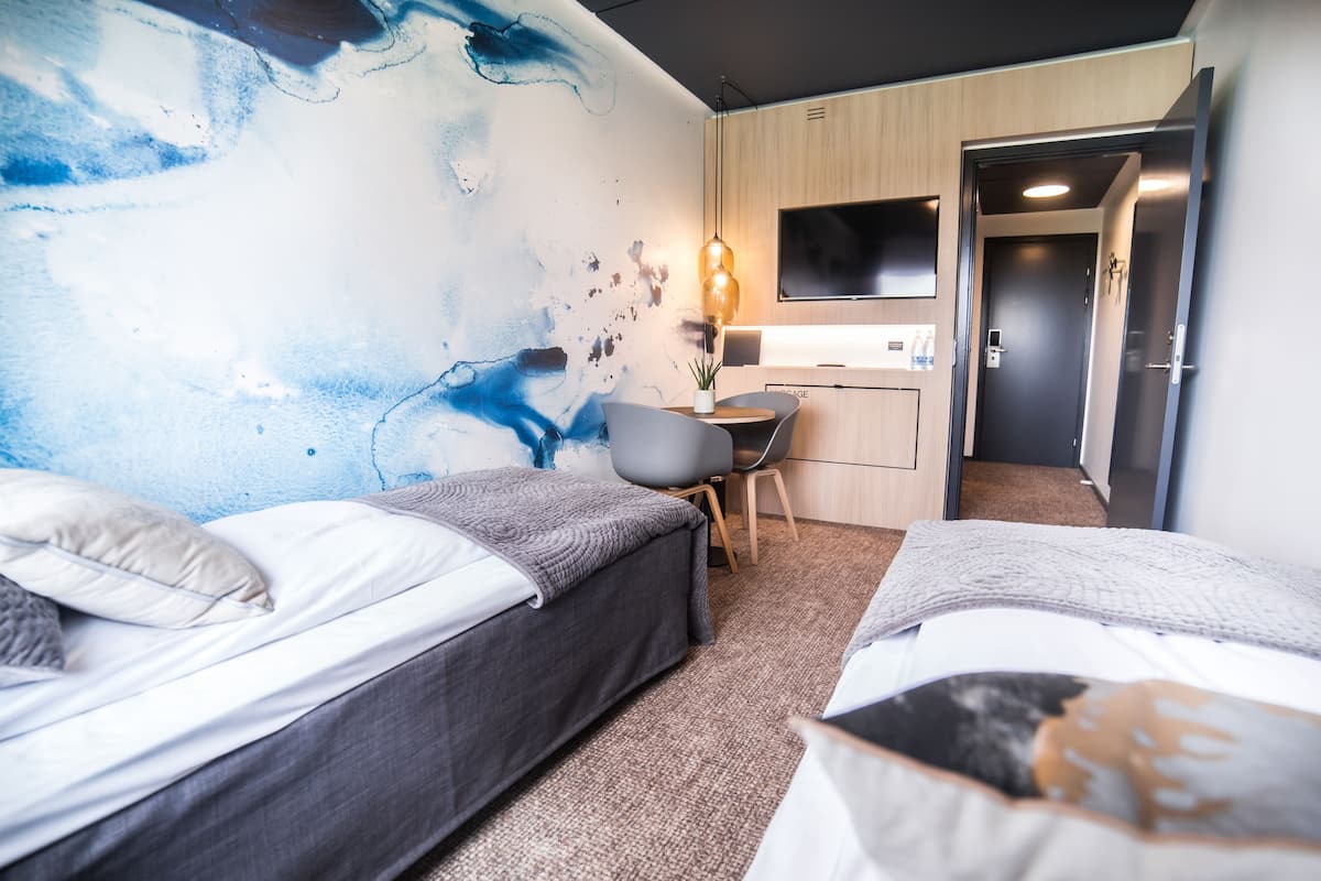 Hotellihuone, jossa on kaksi sänkyä, työpöytä, tuoli, televisio ja sinisellä abstraktilla kuviolla koristeltu seinä.