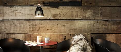 A rustic wooden wall with a hanging pendant light, two black chairs, a small round table with a cup and a menu, and a fluffy white dog.