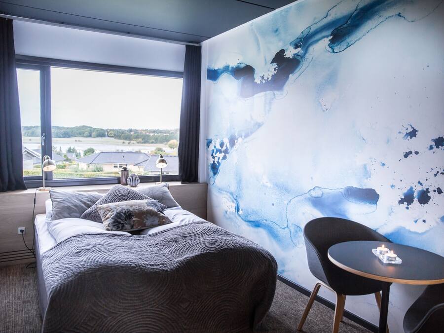 A hotel room with a bed, a small table with a candle, a chair, and a large abstract painting on the wall.