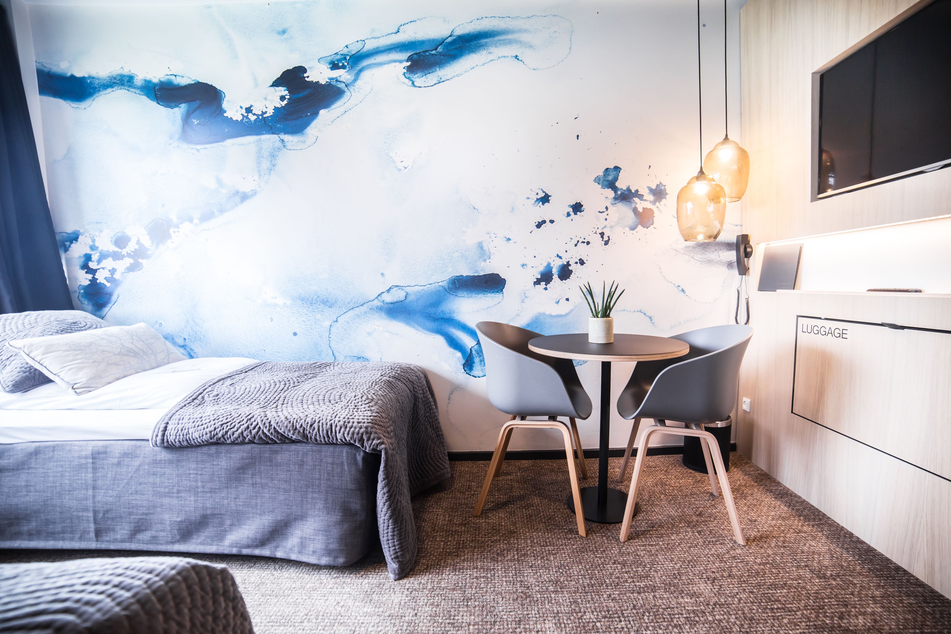 A modern hotel room with a large abstract mural, a bed with grey bedding, a small round table with two chairs, and a luggage storage area.
