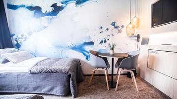A modern hotel room with a large abstract mural, a bed with grey bedding, a small round table with two chairs, and a luggage storage area.