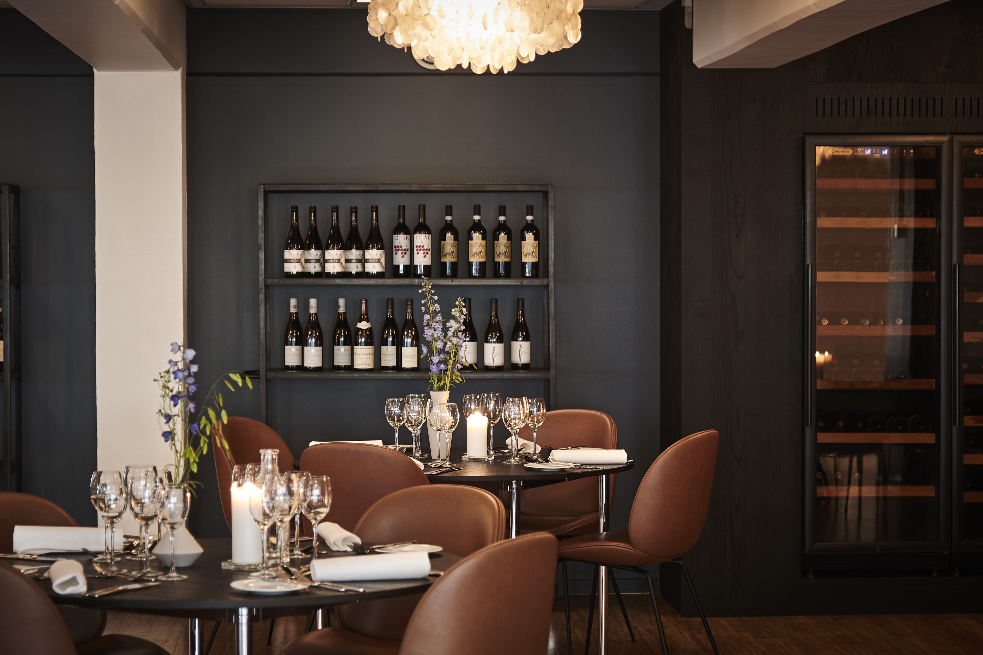 Un comedor con una mesa dispuesta para cuatro personas, rodeada de sillas marrones, una botellera con botellas y un florero decorativo con flores.