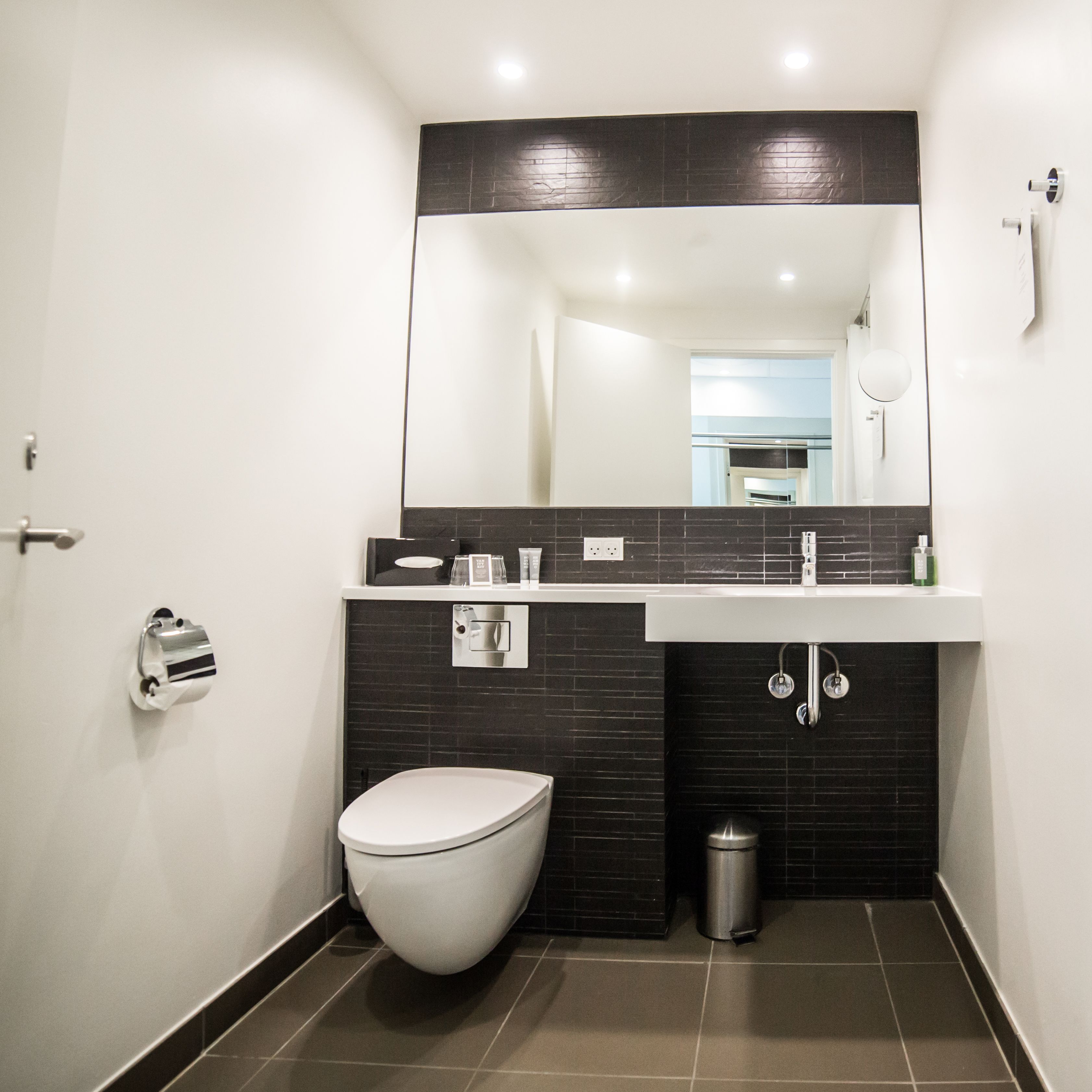 A modern bathroom with a white toilet, a black tiled wall, a white sink, and a mirror above the sink.