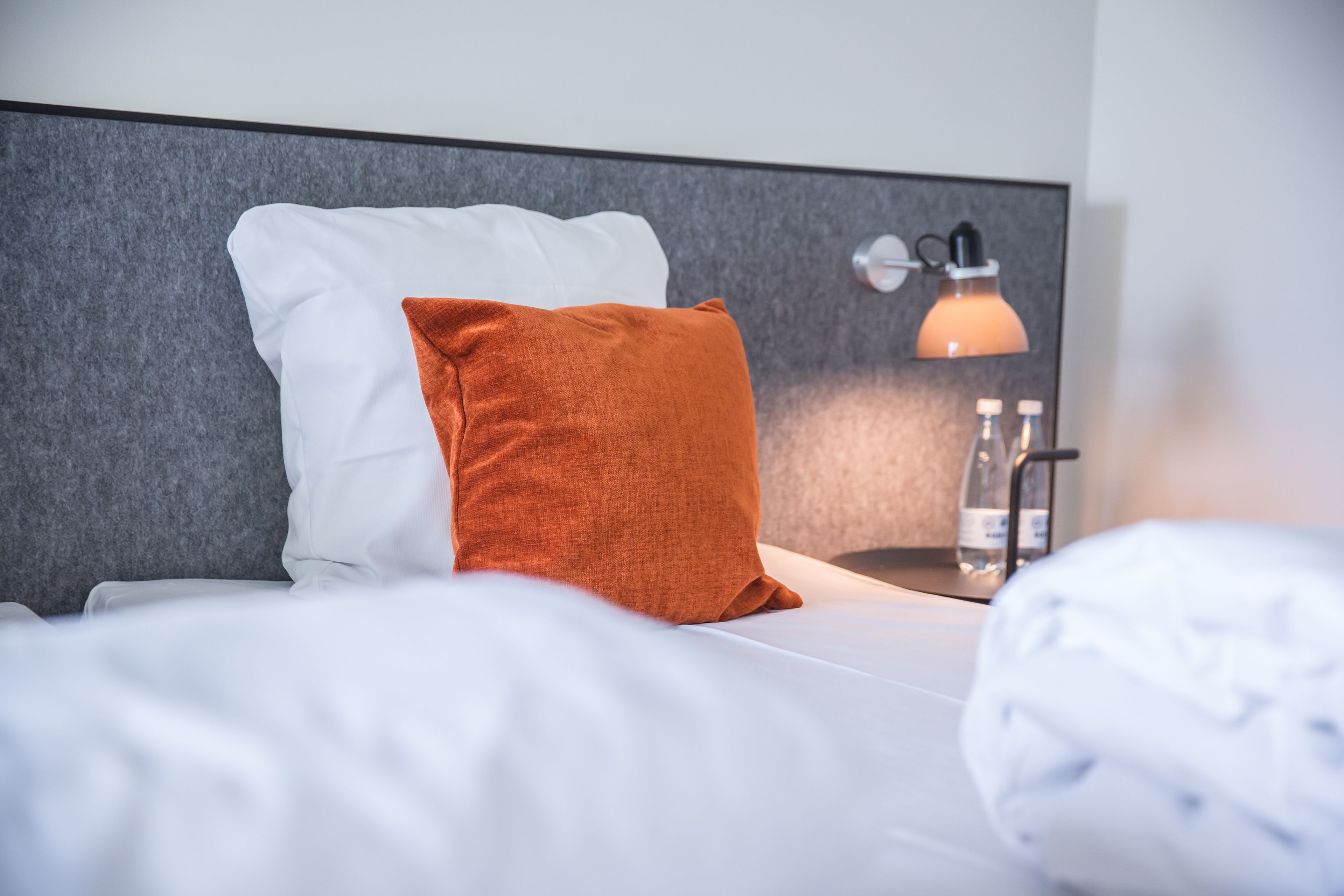 Una cama bien hecha con almohadas blancas y anaranjadas, una mesita de noche con una lámpara y una botella de agua.