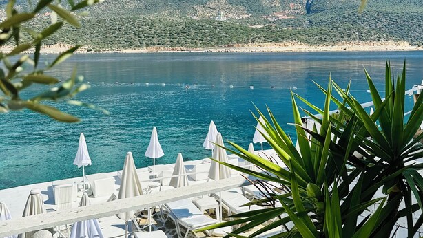 Vista al mar con montañas al fondo, zona de piscina con sillones y sombrillas, y cielo despejado.