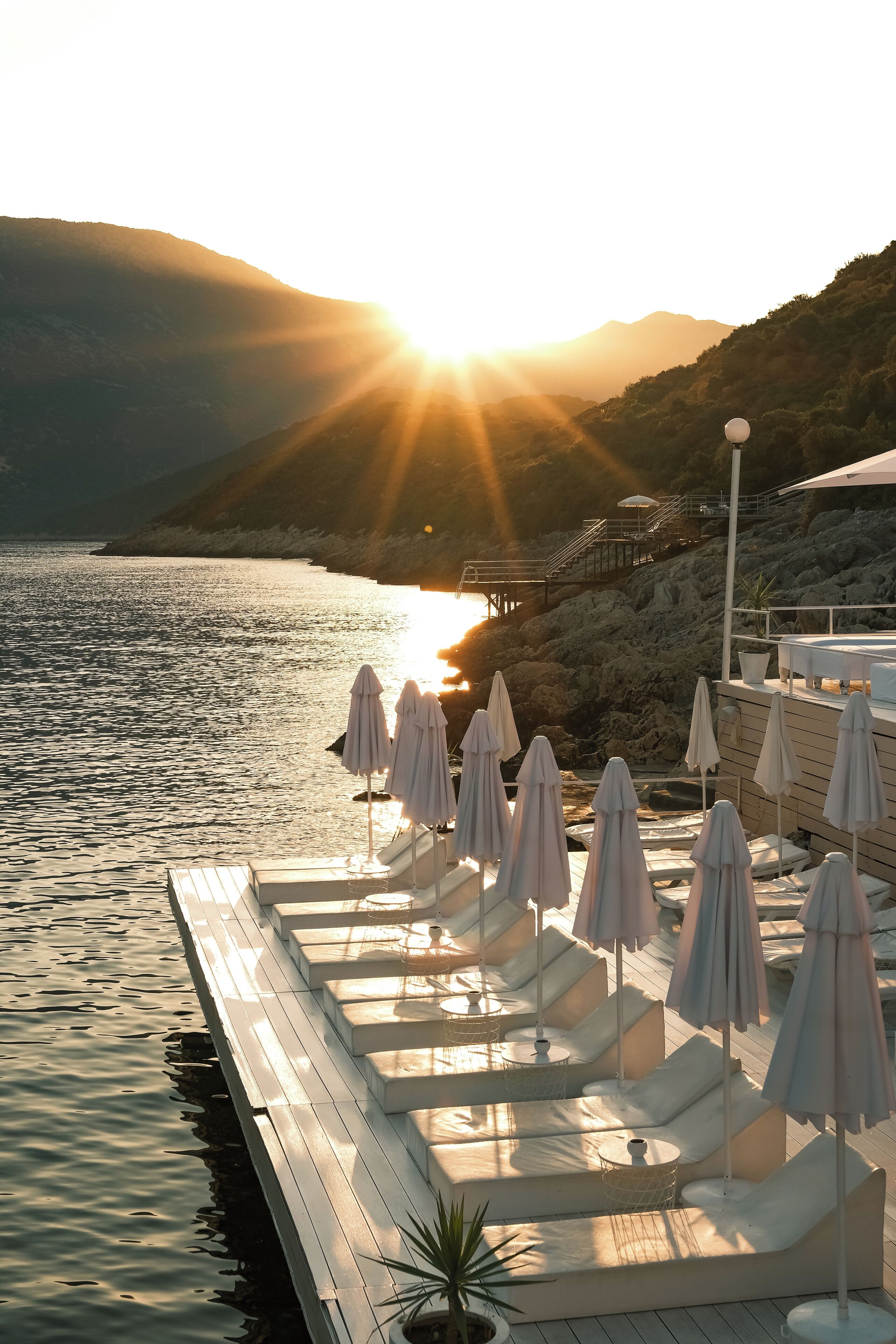 A waterfront deck with lounge chairs and umbrellas, overlooking a body of water with a sunset in the background.