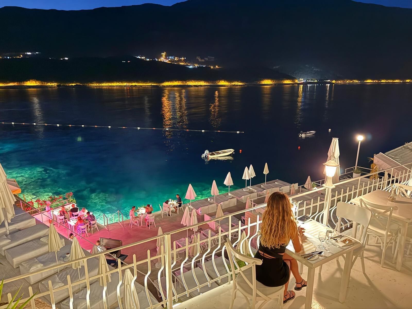 Terraza con mobiliario exterior que da a un lago, un barco y luces de la ciudad a lo lejos.