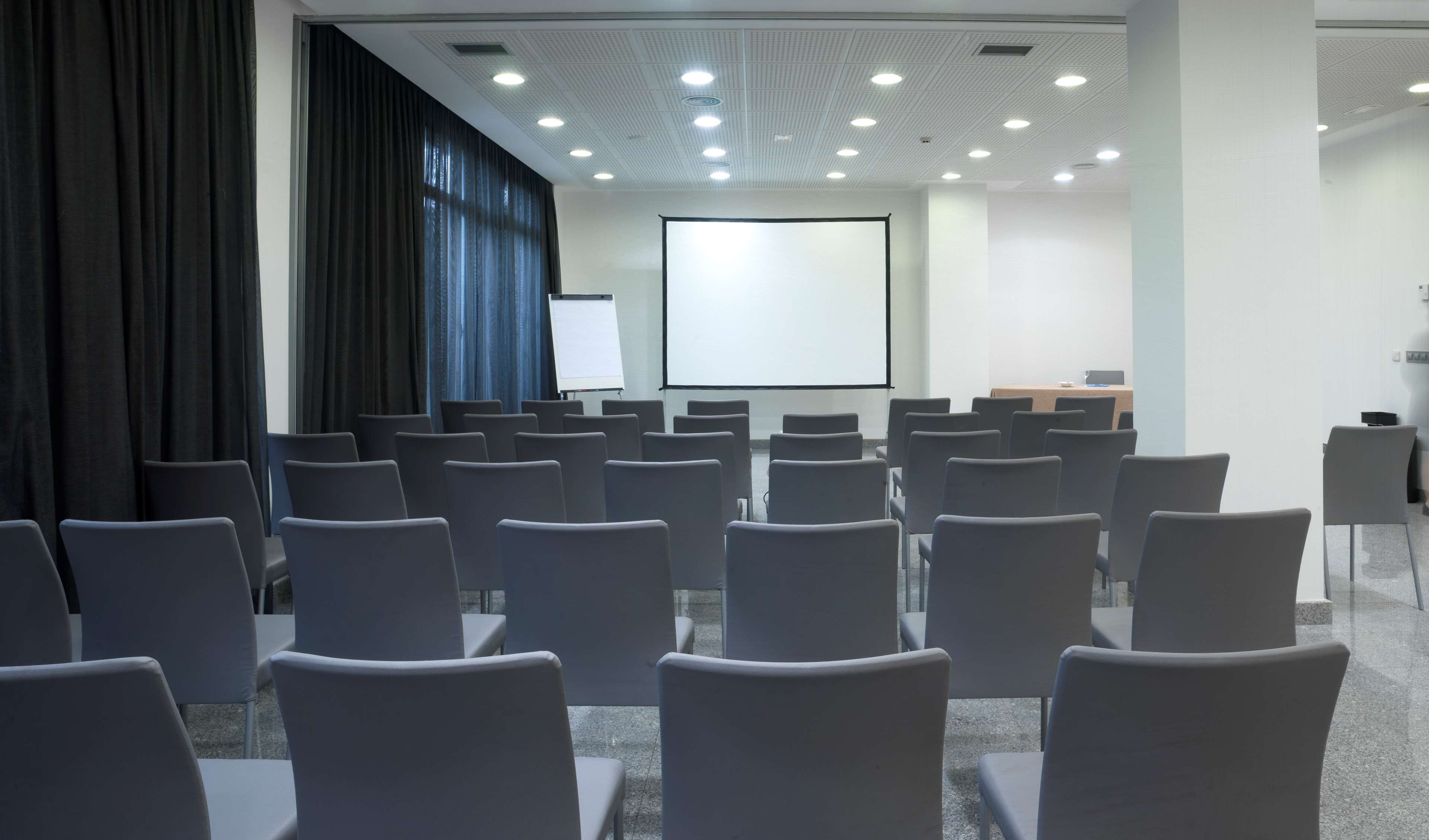 A conference room with rows of chairs facing a projection screen, a whiteboard, and a table with a laptop.