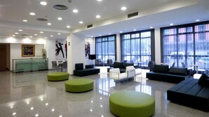 A modern lobby with a glass reception desk, seating areas, and large windows offering views of the parking area.