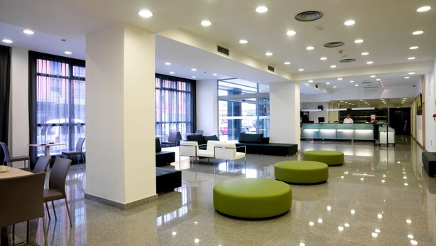 A modern hotel lobby with a reception desk, seating areas, and large windows offering views of the street.